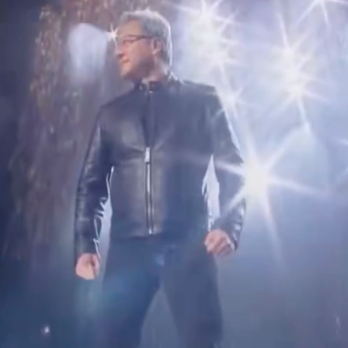 A man in a black leather jacket and dark pants poses dramatically under bright stage lighting with lens flares.