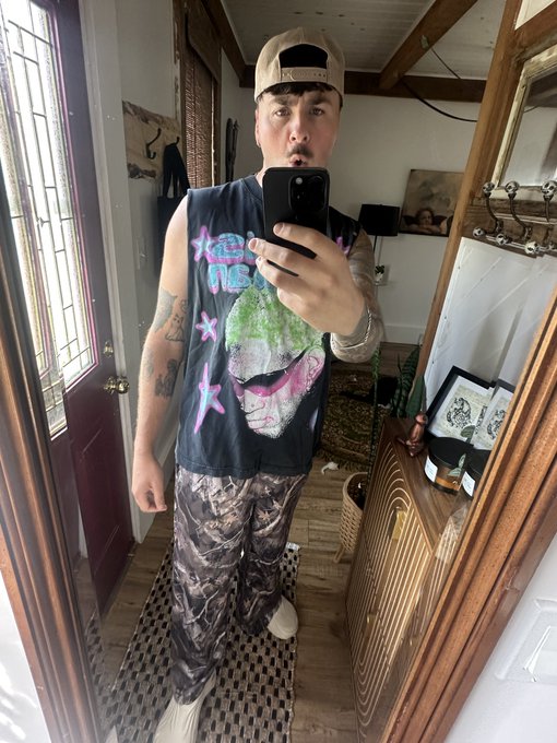 A man takes a mirror selfie in a home entryway, wearing a tan baseball cap, a black sleeveless shirt featuring "JEFF NIPP" text alongside a graphic print, and camouflage pants, with visible tattoos on his arms. The casual, full-length pose complements his friendly "gm good people of crypto twitter" greeting, capturing a relaxed weekend vibe.