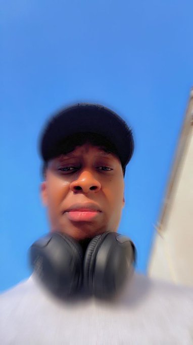 A low-angle selfie of a person wearing a black cap with black headphones around their neck, standing against a bright blue sky with a serious expression.