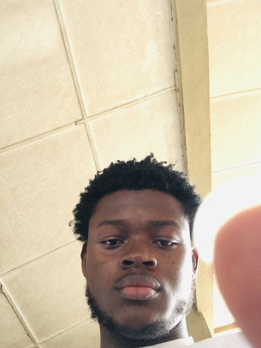 A low-angle selfie of a young Black man with short curly hair and a beard, standing in a tiled room with a finger partially obscuring the camera lens on the right.