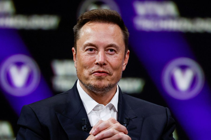 Elon Musk is shown seated at what appears to be a conference panel, wearing a dark suit with his hands clasped together in front of him. The image relates to the breaking news that Musk has publicly endorsed Bitcoin as a superior store of value to cash, reaffirming his personal and corporate (Tesla and SpaceX) holdings of BTC.