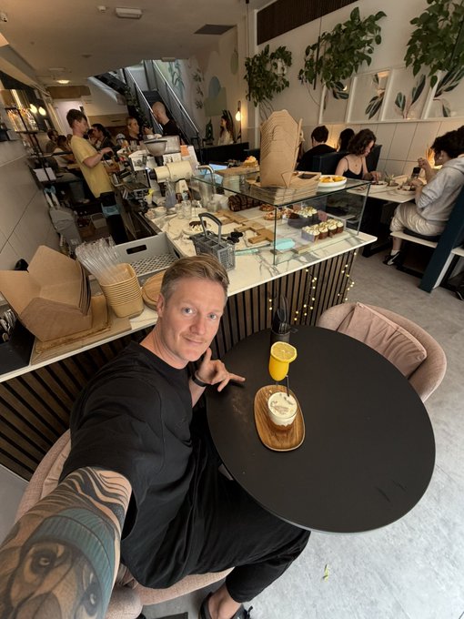 The image features a man sitting at a table with food and drinks, including a cake plate and a cup of coffee or tea. The man has tattoos visible on his arm, adding an artistic touch to the scene. In the background, there are other people seated in chairs around dining tables, suggesting that this is a restaurant setting. A potted plant can be seen near one of the tables, contributing to the ambiance. The image also contains text elements related to travel and adventure experiences, such as "On my way home Stopped at my fav place on route from Barrow to Manchester airport. Love my traditions! Soon Peruvian adventure begins, time to finalize the partners Rok in the cave spaces tomorrow, as usual at 15:00 utc Small cave big vibes, link in BIO." This text seems to be a part of an advertisement or promotional material for a travel-related business or event.