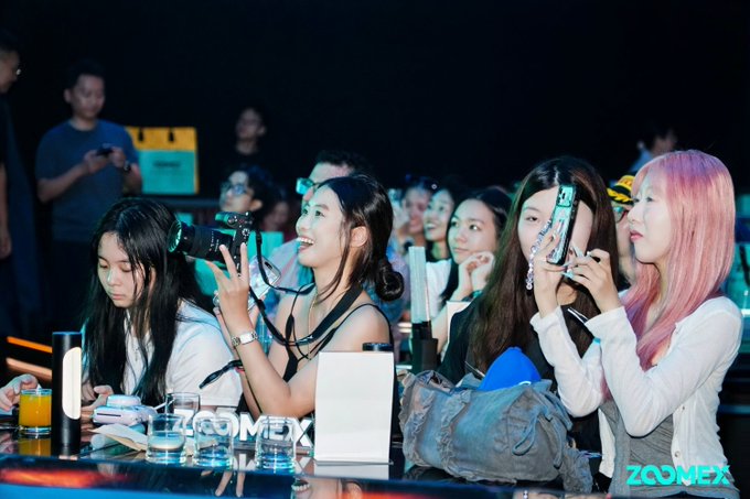 A group of young women at an event with 'ZOOMEX' watermarks, some holding up cameras and phones to take pictures, with one woman having pink hair.