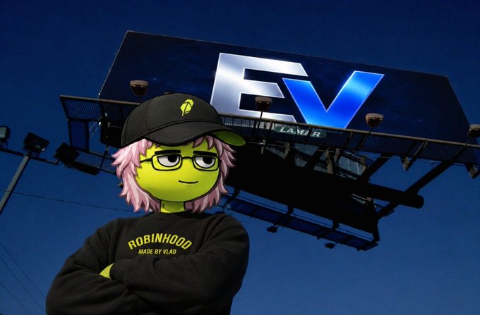 A green-skinned cartoon mascot wearing a black "ROBINHOOD MADE BY VLAD" cap and sweatshirt stands confidently in front of a large EV Law billboard at dusk. The image ties into the giveaway post by using the Robinhood-style branding—suggesting the 20x GTD spots prize as a rewarding, no-cost opportunity to enter Trobiterminal.