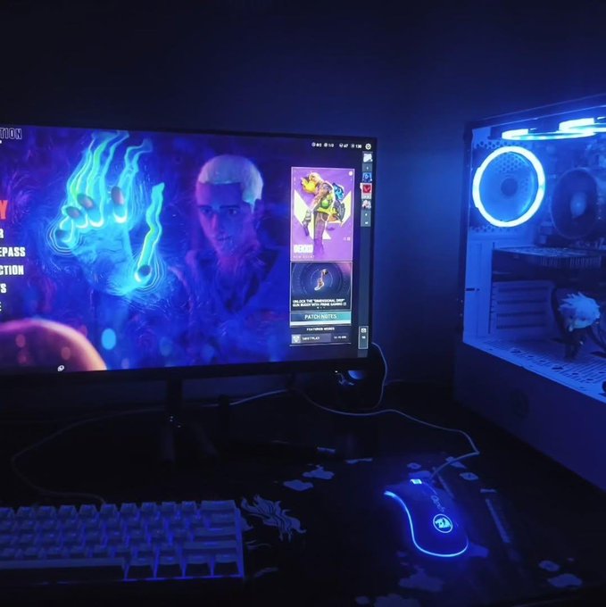 A gaming setup featuring a curved monitor displaying a colorful game scene, an RGB-illuminated PC case with glowing blue fan on the right, and a backlit keyboard and mouse on a dark desk.