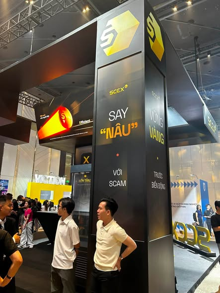 An exhibition hall with a tall black kiosk displaying 'SAY "NẬU" VỚI S - TRỰC TIẾP ĐẾN ĐỒNG' in Vietnamese, with attendees walking around the booth area.