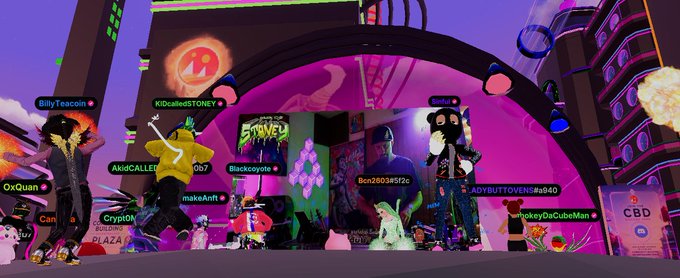 The image shows a group of people standing around an event, possibly at a convention or gathering. The main focus is on a person dressed as "The King" from the video game "Kingdom of Decentraland". This individual is holding a sword in their hand and appears to be in the middle of a performance or speech. There are other people present in the scene, some standing closer to the camera while others are further away. The background features a large archway with lights on it, adding to the atmosphere of the event.