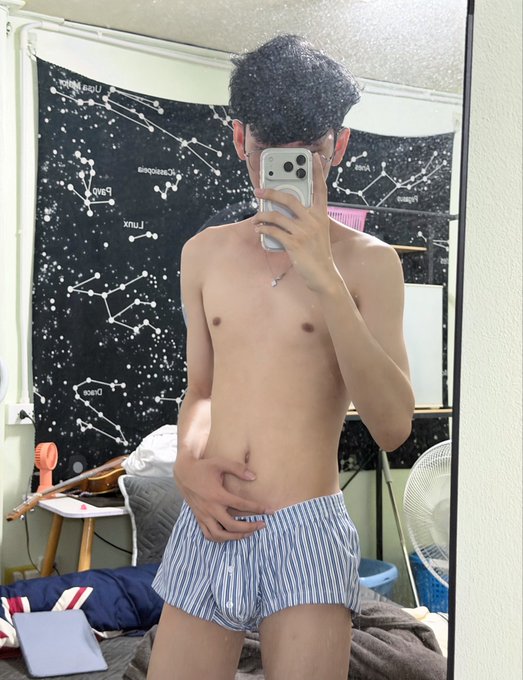A shirtless young man wearing striped underwear takes a mirror selfie with his phone in a bedroom featuring a constellation-themed tapestry on the wall.