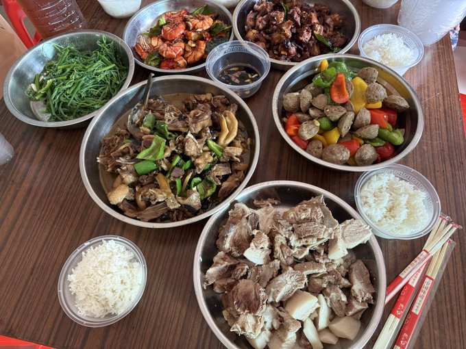 An overhead view of a wooden table laden with a feast of Asian dishes, including braised meats, shrimp, meatballs with bell peppers, greens, and multiple bowls of white rice.