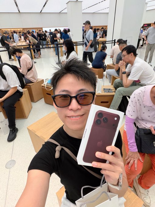 A man takes a selfie at an Apple Store while proudly holding up his newly purchased iPhone 18 Pro Max in its box, with the deep purple color and triple-camera design clearly visible, capturing the excitement of an in-store pickup made using his OKX Card for the 10% cashback deal.