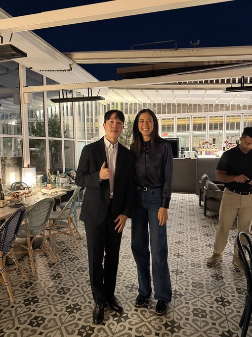 Two people, a man in a dark suit giving a thumbs up and a woman in a satin shirt and jeans, posing for a photo inside what appears to be an upscale restaurant or café with patterned tile flooring.
