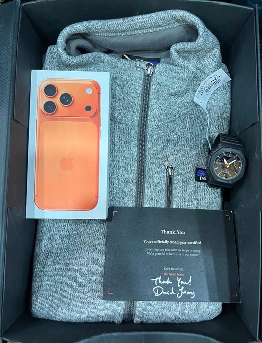 A black gift box contains a neatly folded gray hoodie, an orange iPhone 17 Pro Max in its box, a black G-Shock watch, and a handwritten "Thank You" card from the Tread Fi team, reading "You're officially tread gear-certified. Keep treading." The image showcases the appreciation merch from Tread Fi, aligning with the caption celebrating gratitude and community.