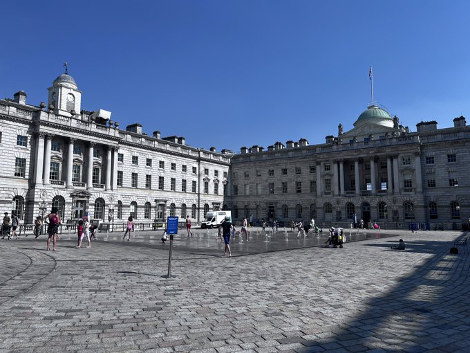 A photograph of Somerset House courtyard in London on a sunny day, showing the grand neoclassical building with its distinctive domed roof, classical columns, and stone facade, with visitors walking around the fountain courtyard area under a clear blue sky.