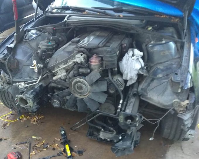 A damaged blue car's engine bay is exposed in what appears to be a garage or salvage yard, with the front end disassembled, parts scattered on the ground, and a rag stuffed near the engine.