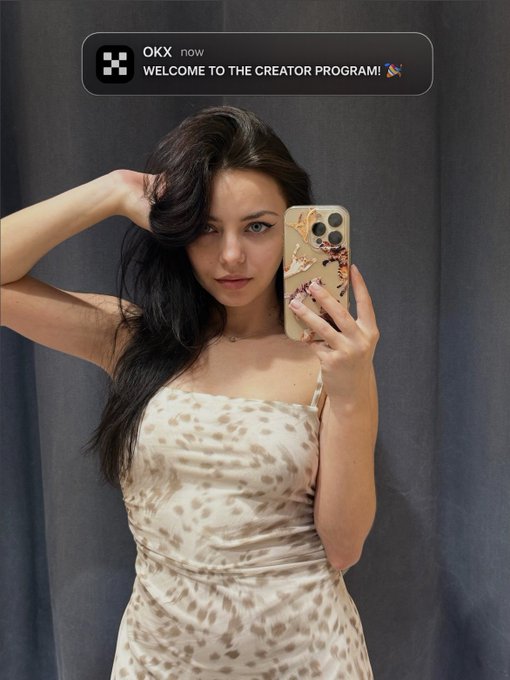 A young woman with long dark hair takes a mirror selfie in a beige leopard-print dress, holding a phone with a decorated case. At the top, a notification reads "OKX now WELCOME TO THE CREATOR PROGRAM! 🎉," celebrating her recent acceptance into the OKX Creator Program, matching her post's excited announcement.