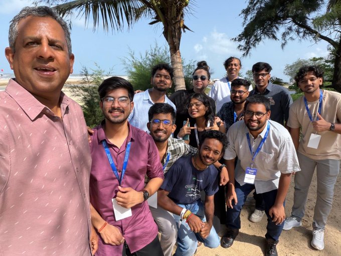 A group photo of a diverse group of people, including a man in a pink shirt taking a selfie, posing outdoors near a beach with palm trees.