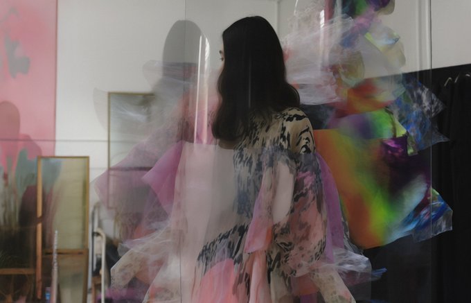A mannequin wearing a flowing garment is reflected on a glass panel, surrounded by vibrant pink, purple, and iridescent fabrics, as if saying "Gm" with a soft, artistic morning glow.