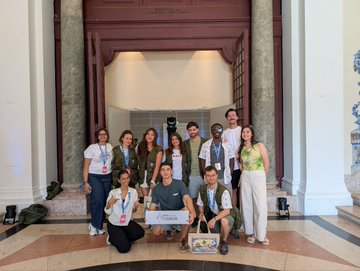 A group photo of people posing in front of an ornate building entrance, with two people in the front holding boxes/packages, one of which appears to be a Logitech product.