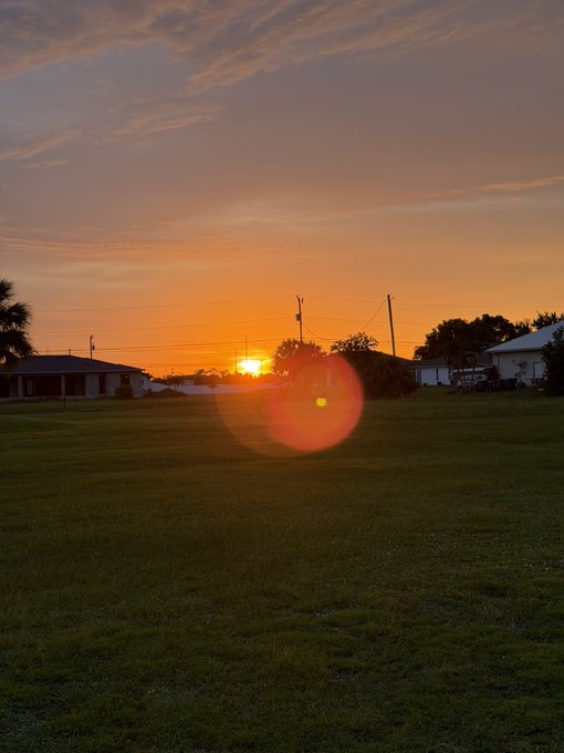 A scenic photograph of a sunset over a grassy field with houses and power lines in the distance, featuring a prominent orange lens flare in the center.