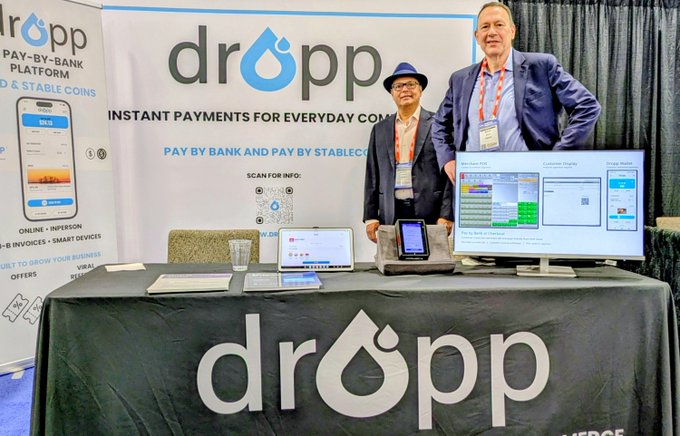 Two men stand behind a promotional booth for 'dropp', a pay-by-bank and stablecoin platform, displaying their services with monitors and a tablet device.