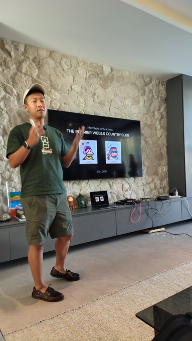 The image shows a man standing in front of a television screen, which displays two different images side by side. One image features a cartoon character with a hat, while the other depicts a cartoon character wearing a baseball cap. The man appears to be giving a presentation or lecture on these two characters and their differences.
