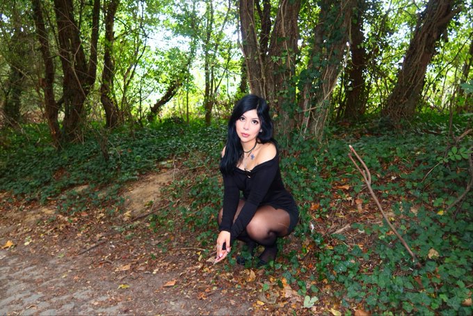 A young woman with long black hair crouches on a dirt path in a lush, leafy woodland, dressed in a black top and tights, holding a small pink object. The scene captures a casual outdoor photo that fits the "cute lil digital pics" caption, showcasing a soft, nature-inspired portrait.
