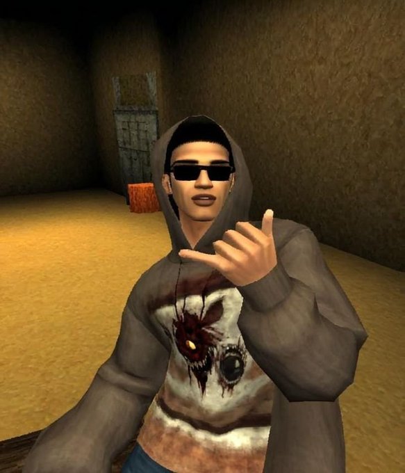 The image features a man wearing sunglasses and a hoodie with a skull design on it. He is sitting down and making a peace sign with his fingers. The background of the image shows an empty room or hallway, which could be part of a video game setting. There are also two boxes visible in the scene, one located near the center-left side of the image and another towards the right edge.