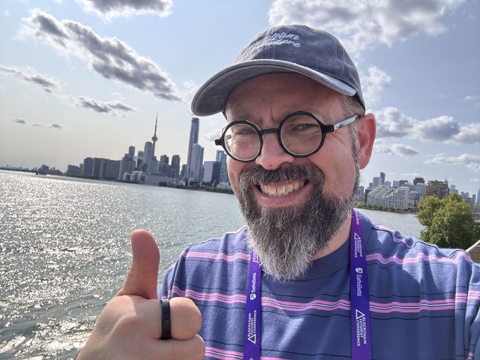 A selfie of a bearded man wearing a Disrupt cap, glasses, and lanyard, giving a thumbs-up with the Toronto skyline visible across the waterfront.