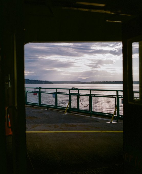 A view from inside a ferry or boat looking out through an open doorway onto the car deck, showing a metal railing, calm water, and a cloudy sky with distant hills on the horizon.