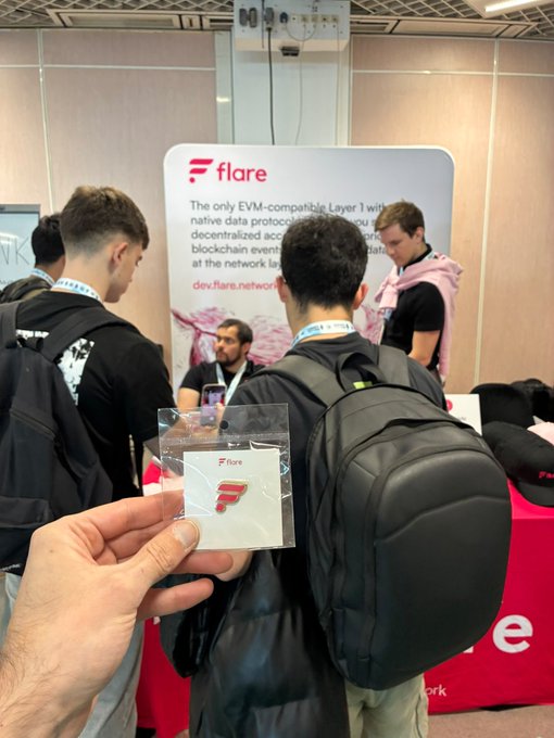 The image shows a group of people gathered around a table at an event, with one person holding up a small white card or sticker that says "Flare" on it. The background includes a banner promoting the company's name and logo. The attendees are engaged in conversation and enjoying themselves, creating a lively atmosphere for the event.