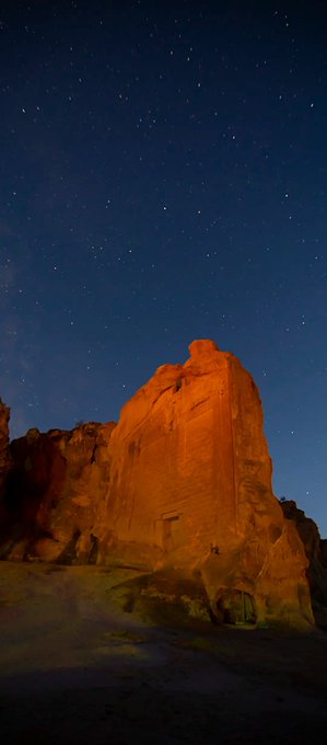 Nighttime photograph of a large rock formation or cliff face illuminated by warm light, with a starry night sky and the Milky Way visible above.