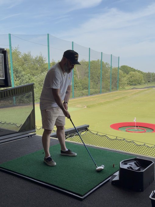 A bearded man wearing a black cap, gray t-shirt, and khaki shorts plays golf at a driving range, swinging a club toward a target on the green.