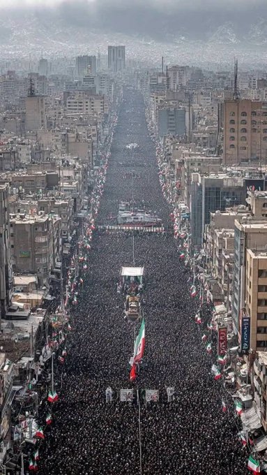 An aerial view of a massive crowd filling a wide avenue in an Iranian city, with Iranian flags visible and the crowd extending as far as the eye can see.