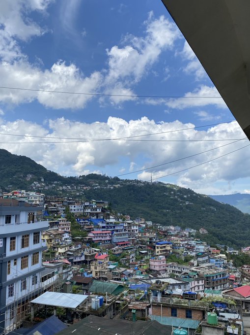 A daytime view shows a densely populated hillside town with colorful buildings nestled in a mountainous landscape under a bright blue sky with scattered clouds.