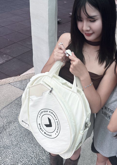 A young woman smiles as she looks into a cream-colored backpack featuring the text "BIRDSDAY FINANCE OFFICIAL BIRDS Wallet" displayed in a circular logo. The post celebrates Bitget Wallet's 100 million users and 8th anniversary, highlighting the branded bag and bracelet as anniversary gifts alongside promotion of their 3% cashback U-card feature.