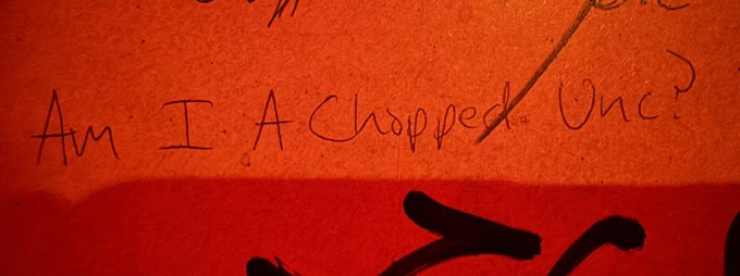 Handwritten text in red ink on an orange paper surface reads "Am I A Chopped One?" with a black painted shape below.