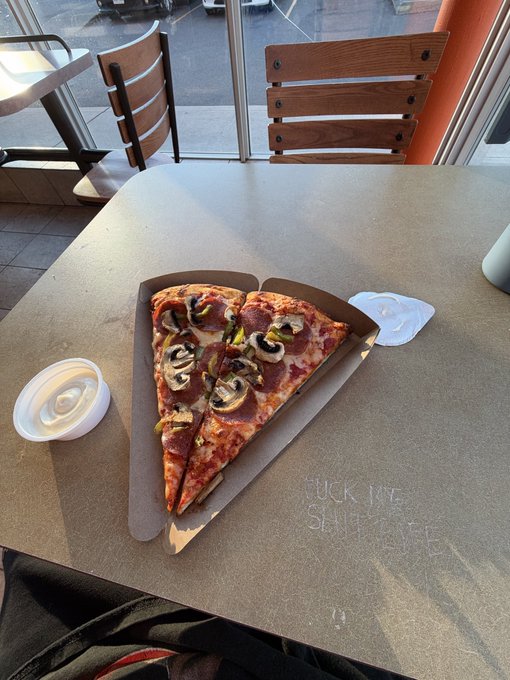 Two slices of pizza topped with mushrooms and meat on a paper tray, served on a table with a small container of white sauce.