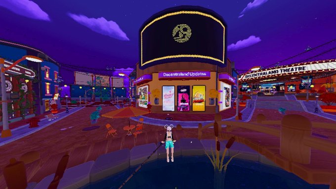 A virtual 3D environment showing a colorful plaza with a central building displaying a logo, surrounded by various storefronts including 'DREAMALAMI THEATRE' and 'Decentraland Updates'.