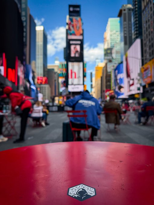 A bright red table with an MLB-logo sticker anchors the foreground of this bustling Times Square scene, where a person in a blue "SUPERHERO" jacket sits facing towering billboards under a blue sky—setting the stage for the epic Saturday ahead.