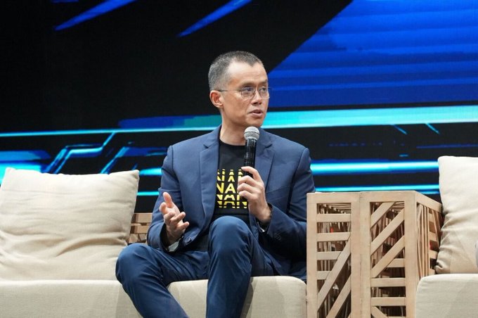 Man wearing glasses and a dark suit jacket over a black t-shirt with yellow text, speaking into a microphone while seated on stage at what appears to be a conference.
