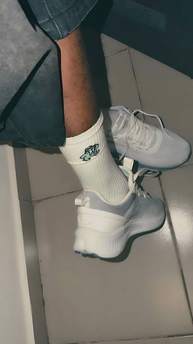 A person wearing white sneakers, white socks with a green Nike logo, and denim pants stands on a tiled floor.