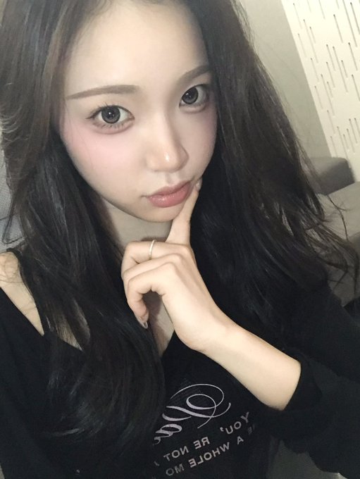 A close-up selfie of a young woman with long dark hair, dramatic eye contacts, posing with her hand near her face while wearing a black shirt with white text.