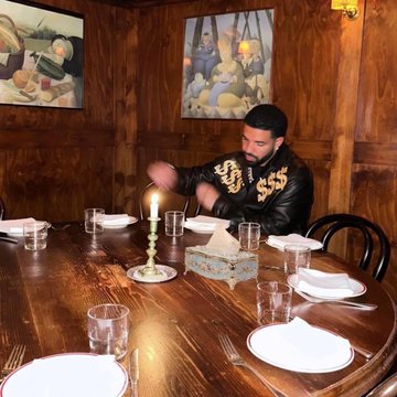 The image shows a man sitting at a table with glasses of water, wine glasses, and cups placed around him. He is wearing a black leather jacket and has his hands up in front of him. The background features paintings hanging on the wall above the dining table. There are also two other people present in the scene, one standing near the left side of the image and another person sitting at the far right edge of the frame.