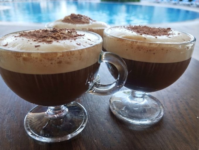 Two glass mugs filled with dark coffee topped with a thick layer of frothy foam and a sprinkle of cocoa powder, sitting on a wooden table near a swimming pool.