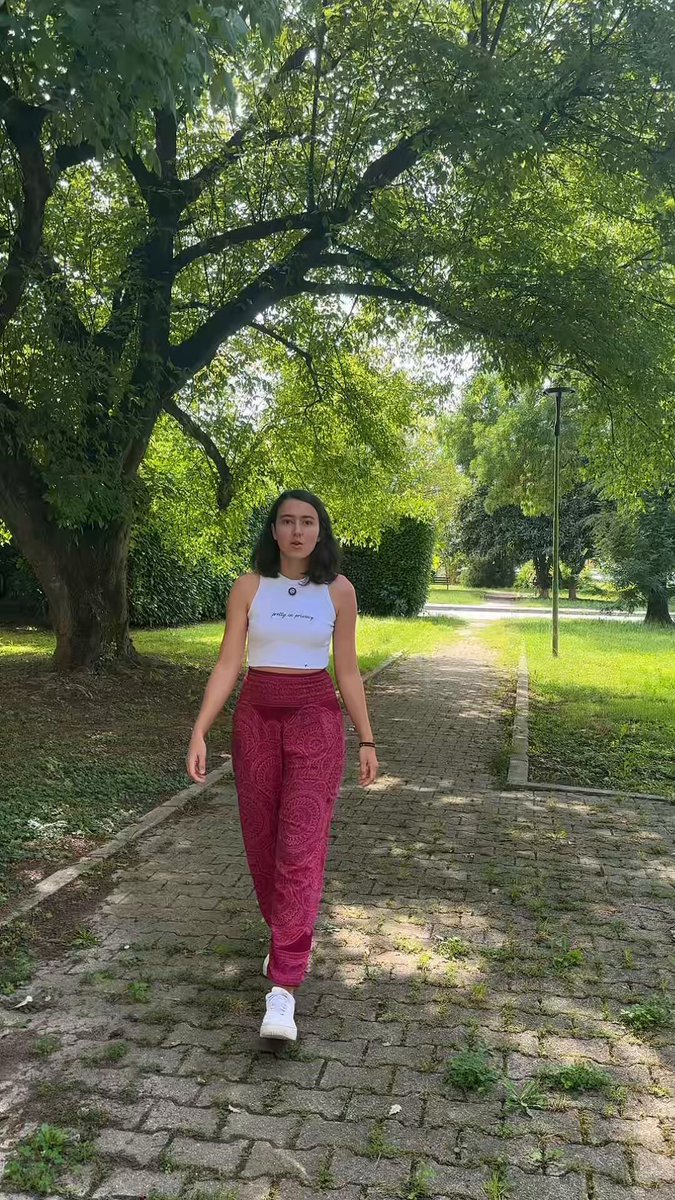 The image features a woman walking down a path wearing red pants and a white tank top. She appears to be enjoying her walk through an outdoor setting surrounded by trees. The photo has been edited with a filter that makes the colors appear more vibrant, giving the scene a lively atmosphere.