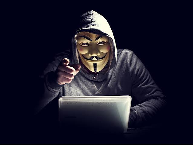 A hooded figure wearing a Guy Fawkes mask points at the camera from behind a glowing laptop in a dark setting, symbolizing anonymous cyber threats and hacking. The image visually reinforces the post's warning about looming quantum computing threats that could exploit exposed public keys and break Bitcoin's encryption within years.