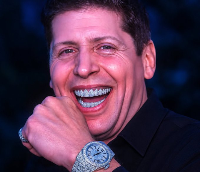 A close-up portrait of a smiling man with diamond-encrusted teeth and a diamond-covered wristwatch, wearing a black shirt.