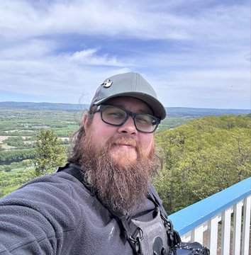 A bearded man wearing glasses and a gray cap takes a selfie overlooking a scenic landscape of green trees and hills under a blue sky.
