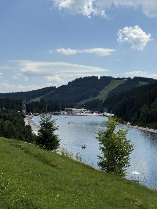 A scenic view of a lake surrounded by forested hills with a ski slope visible on the mountainside; boats are on the water and green grass fills the foreground.
