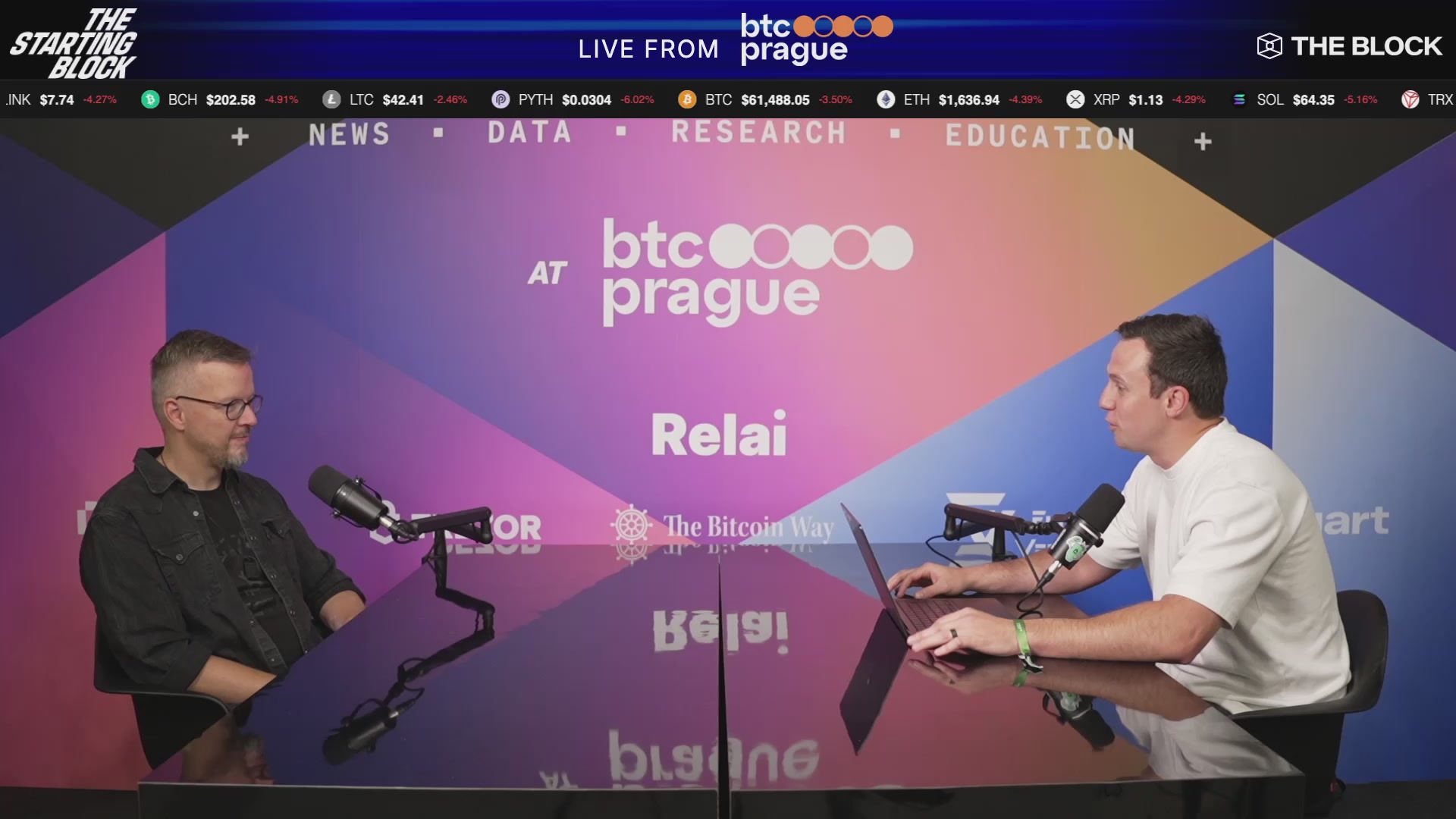 The image shows a news broadcast featuring two men sitting at a table with microphones, both of which appear to be computer monitors or laptops. They are appearing on a television screen placed behind them. The hosts seem focused on their discussion topic. There is also text visible in the photo that reads "Live from BTC Prague - Day 1". This might indicate that this live broadcast is covering a newly released cryptocurrency event, and it could possibly explain why there's a difference between day one of the podcast being discussed or the fact that there are two men participating in the discussion.